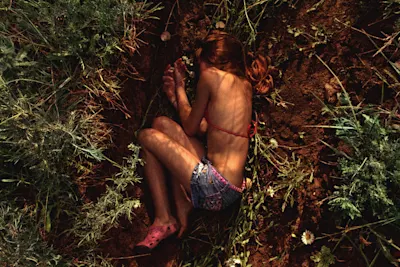 Exhibition Circulation(s) 2024 - Focus Ukraine Woman in shorts and bikini top lying curled up on the ground, surrounded by grass and flowers.