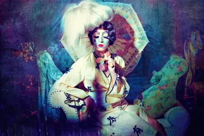Jamari Lior – Fotokunst Frau in traditionellem Outfit mit Schirm auf Sessel.