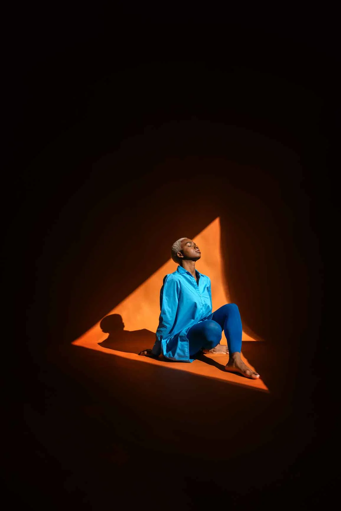 Una persona vestida de azul está sentada sobre un triángulo iluminado en naranja sobre un fondo oscuro y mira hacia arriba. Una persona vestida de azul está sentada sobre un triángulo iluminado en naranja sobre un fondo oscuro y mira hacia arriba.