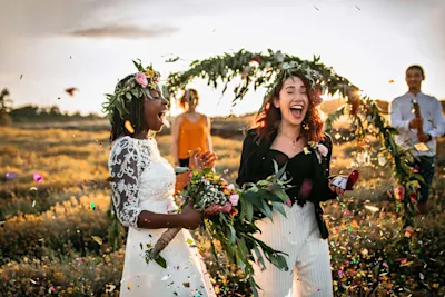 Bride in front of a wedding arbour A bride in a white lace dress holding a bouquet, and a woman in a black jacket and white pants, standing in front of a flower-decorated arch outdoors at sunset, with confetti in the background.