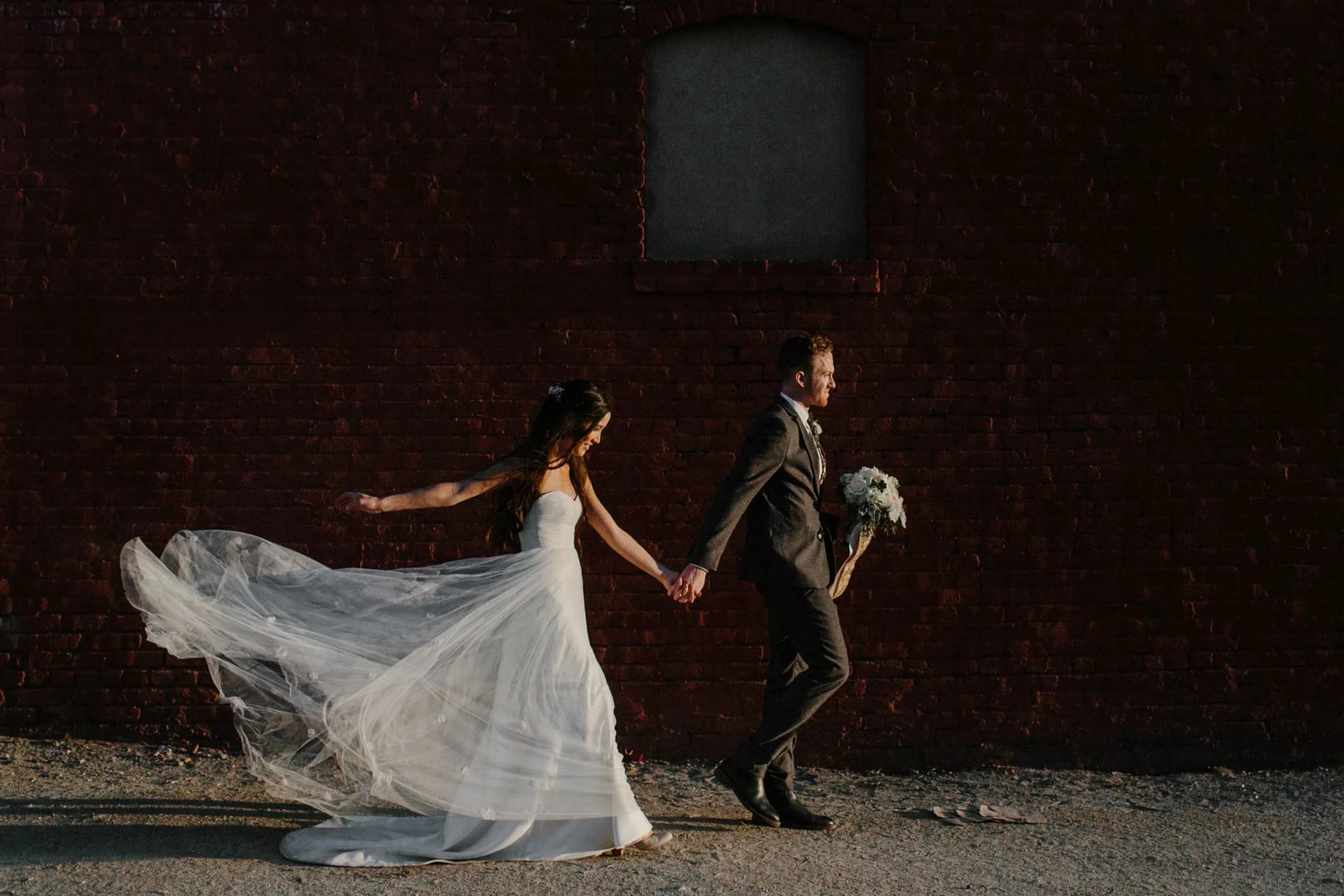 Bride and groom A bride in a flowing white dress holds the hand of the groom, who is wearing a suit and carrying a bouquet, in front of a dark brick wall.