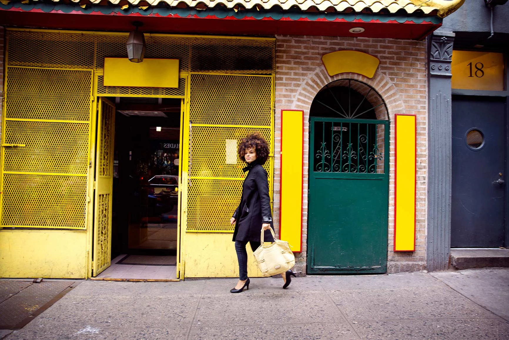 Woman in front of a building with a green and yellow door A woman with curly hair, dressed in black, is carrying a light-colored bag and walking past a building with yellow and green doors.