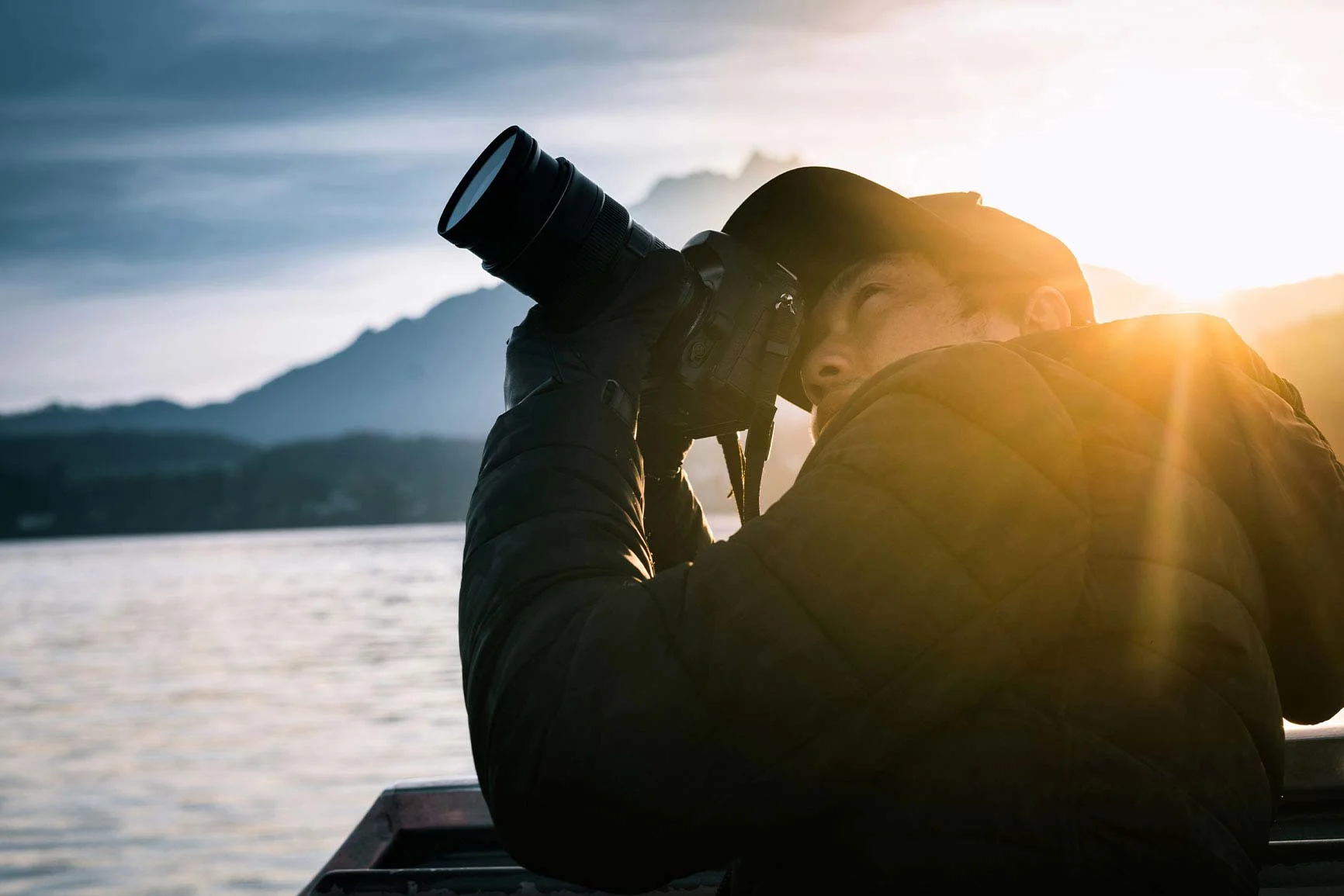 A man with a camera on a boat A man with a camera on a boat takes a photo at sunset over a lake with mountains in the background.