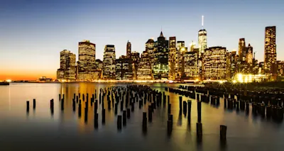 Skyline bei Nacht Beleuchtete Skyline einer Stadt bei Nacht, davor Holzpfähle im Wasser.