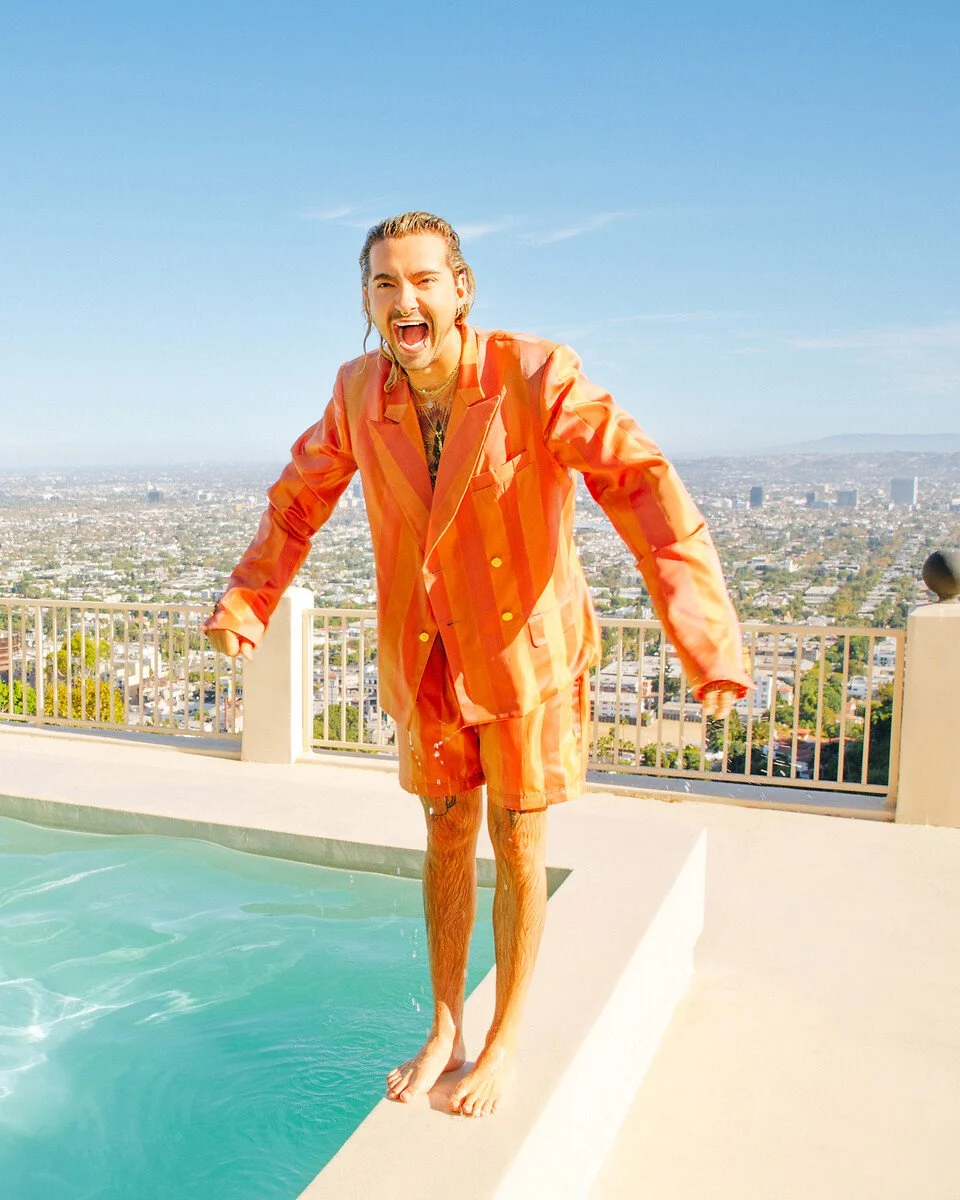 Bill Kaulitz lacht an einem Pool mit Skyline-Blick Ein Mann in einem orange gestreiften Anzug lacht an einem Pool auf einer Dachterrasse mit einem weiten Blick auf die Skyline der Stadt unter einem klaren blauen Himmel.