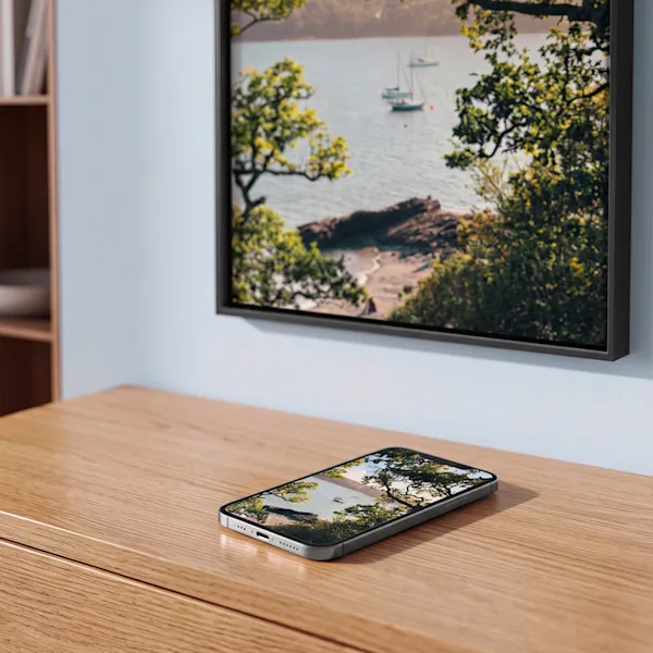 Smartphone-Foto als Wanddruck Ein Smartphone auf einem Holzschreibtisch, das eine Küstenszene anzeigt, passend zu einem gerahmten Wandbild mit Segelbooten und Bäumen darüber.