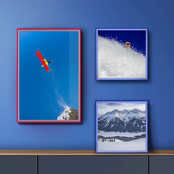 Wall design with colorful Pop Art frames Three wall prints of different sizes are arranged creatively on a blue wall above a sideboard. The pictures are encased in striking, colorful frames in red and blue. The motifs showcase dynamic winter sports scenes: a ski jumper in mid-air, a freeskiier in deep powder, and a wintry mountain panorama.