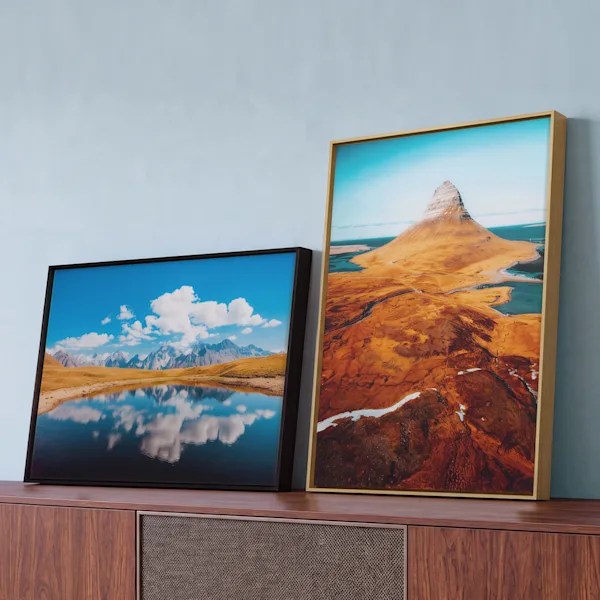 „WhiteWall Selected“ gerahmte Fotoabzüge Zwei gerahmte Landschaftsdrucke an einer hellblauen Wand: eine Spiegelung eines Bergsees in einem schwarzen Rahmen und eine Luftaufnahme einer Berglandschaft in einem hellen Holzrahmen.
