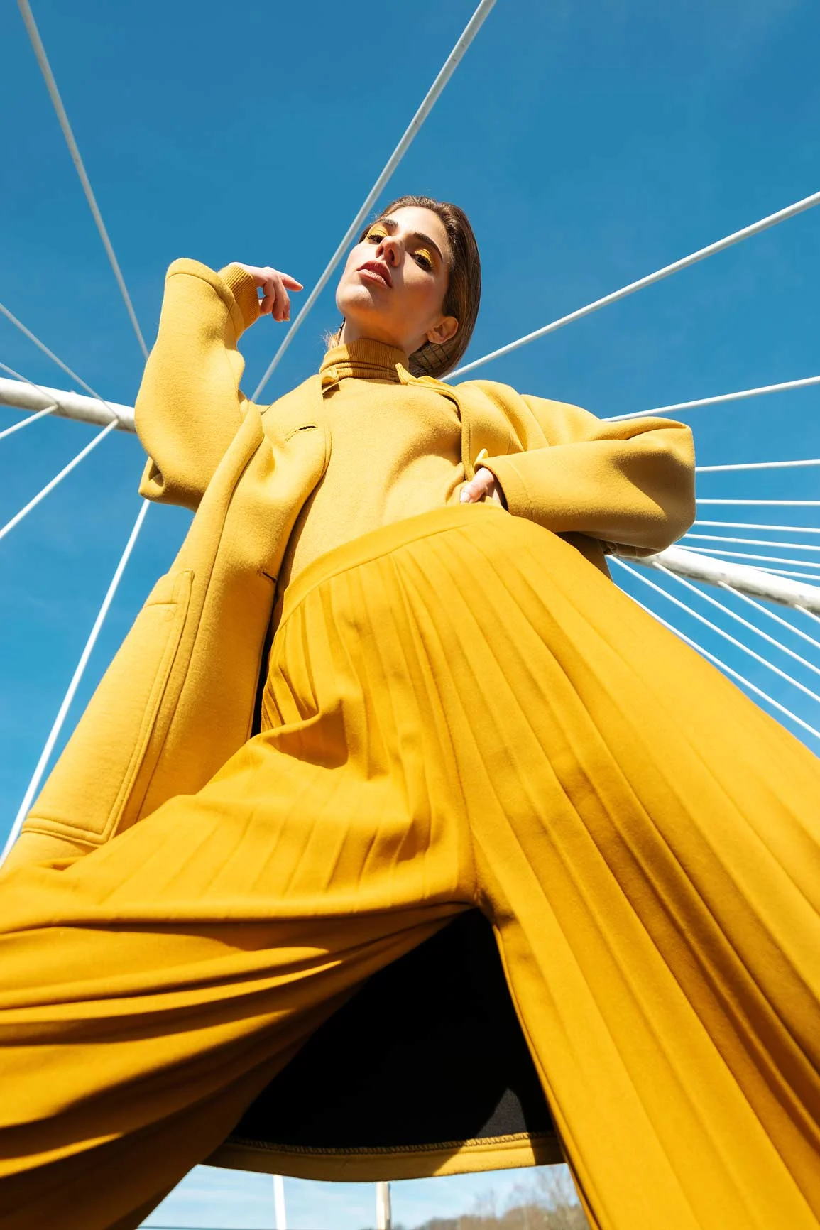 Mujer con abrigo amarillo bajo un cielo azul con cables de acero, fotografiada desde abajo. Mujer con abrigo amarillo bajo un cielo azul con cables de acero, fotografiada desde abajo.