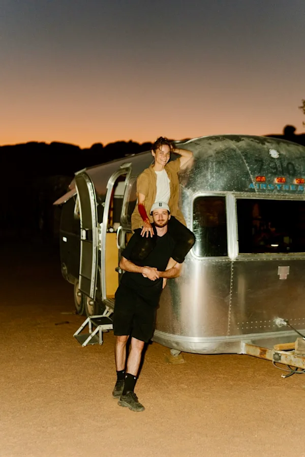 Portrait de Thylacine et Cécile Chabert devant le studio mobile Un portrait pris au crépuscule dans le désert namibien. Le musicien Thylacine se tient au premier plan devant une caravane Airstream argentée, portant Cécile Chabert sur ses épaules. Tous deux sourient à l'objectif. Thylacine porte un t-shirt noir, un short sombre et une casquette claire ; Cécile porte une chemise marron clair sur un haut blanc. L'arrière-plan montre les silhouettes sombres du paysage rocheux sous un ciel de soirée orange et bleu.