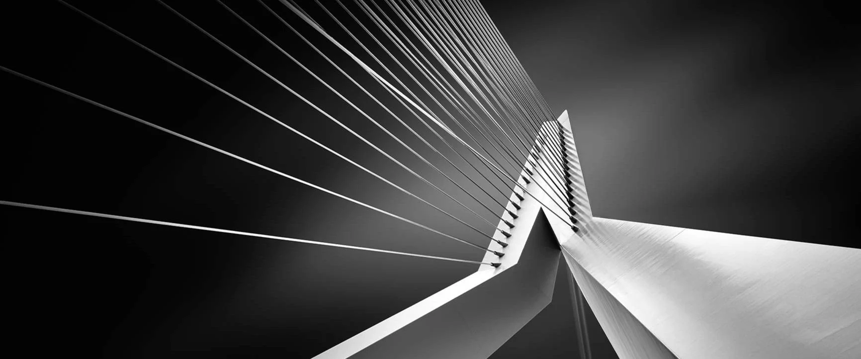 A black-and-white photograph of a modern suspension bridge taken from a low angle, with the suspension cables visible.
