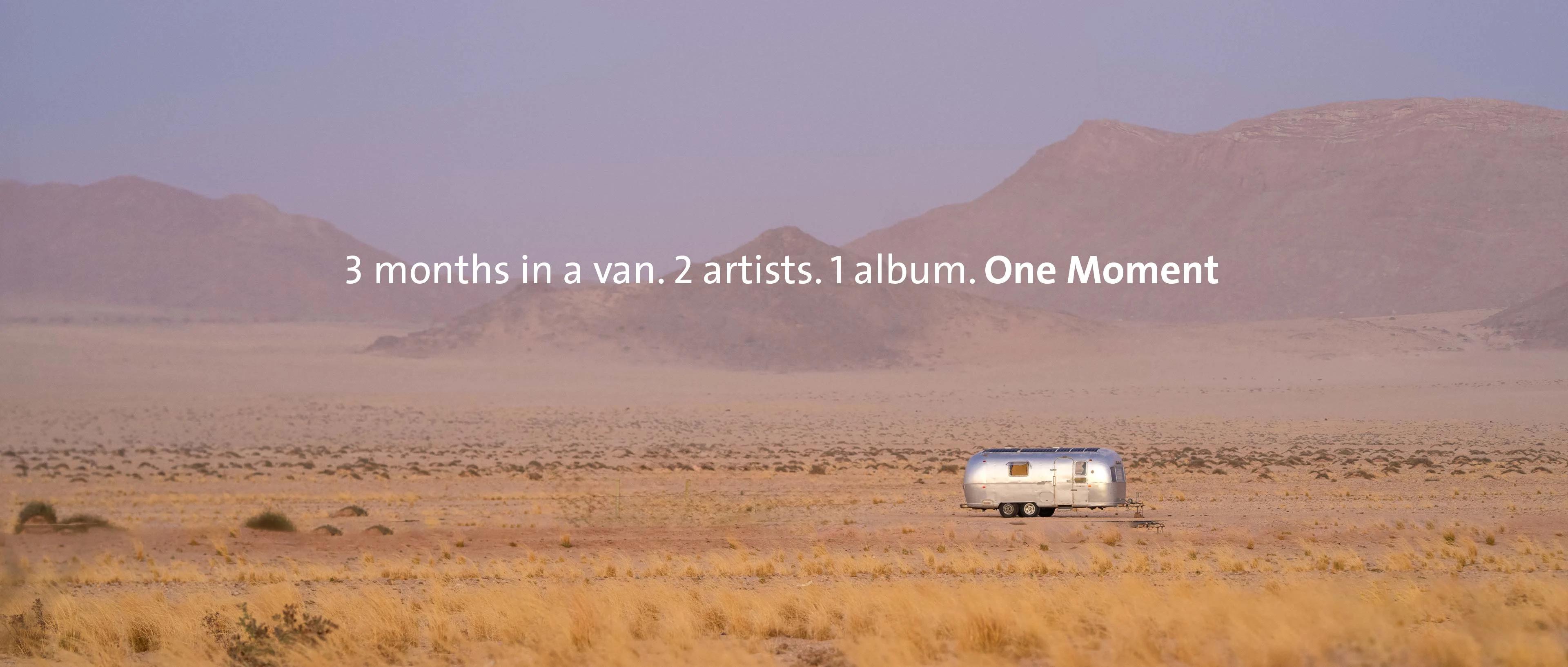 A wide panoramic shot of the Namibian desert in diffused purple light. In the middle of the sandy plain stands a silver Airstream trailer in front of a gentle mountain range. Centered in the image is the text: "3 months in a van. 2 artists. 1 album. One Moment".