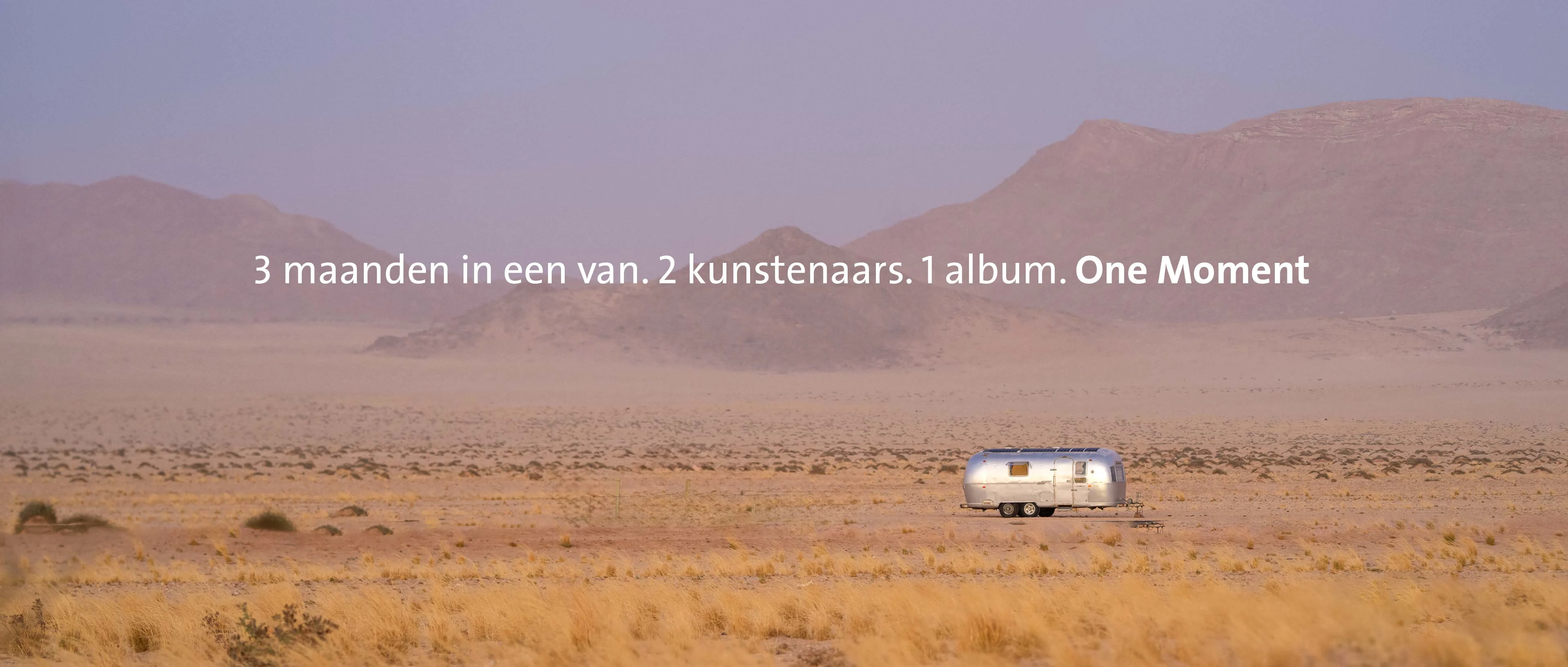 Een weidse panoramische opname van de Namibische woestijn in diffuus paars licht. Midden in de zandvlakte staat een zilveren Airstream-caravan voor een glooiende bergketen. Centraal in het beeld staat de tekst: "3 maanden in een busje. 2 artiesten. 1 album. One Moment".