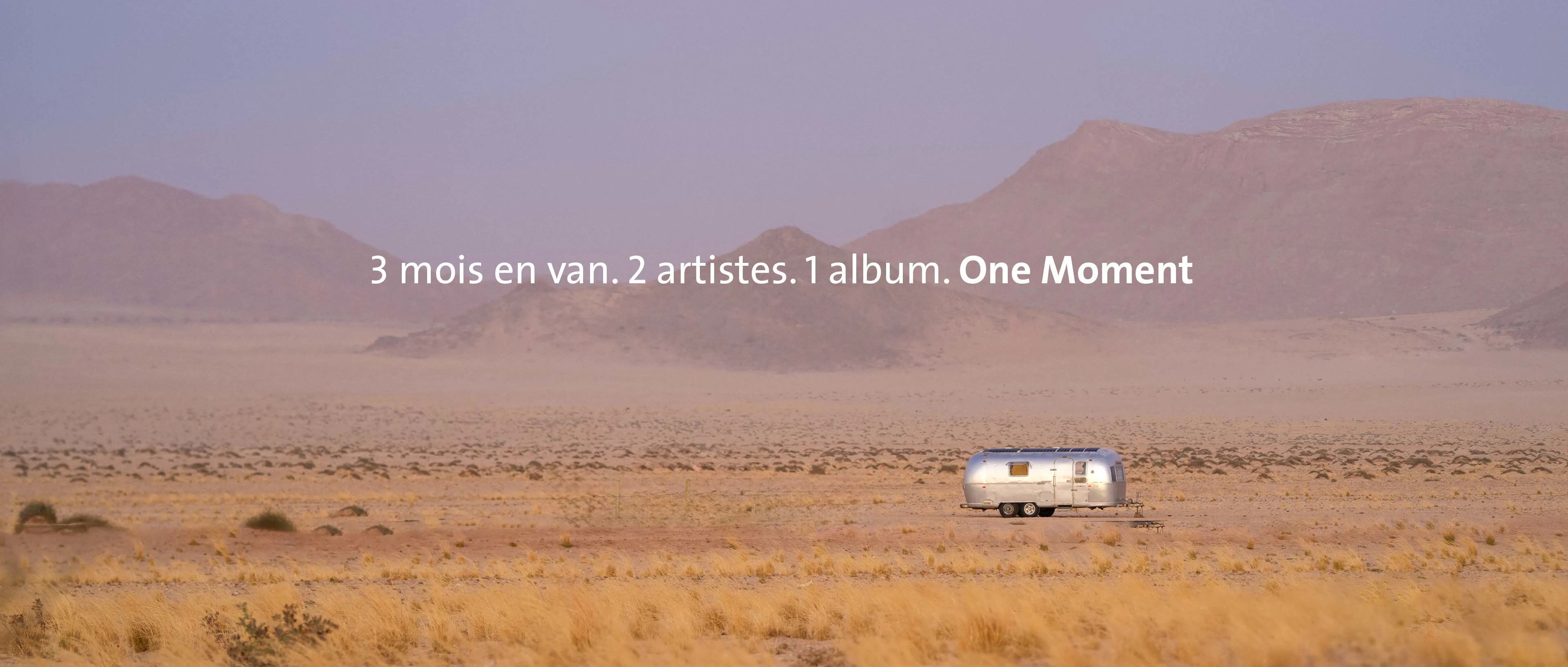 Une large vue panoramique du désert namibien dans une lumière violette diffuse. Au milieu de la plaine sablonneuse se trouve une caravane Airstream argentée devant une douce chaîne de montagnes. Au centre de l'image figure le texte : "3 mois dans un van. 2 artistes. 1 album. One Moment".