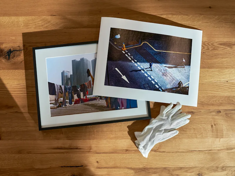 Fine Art Print 4 Two fine art prints on wooden surface, one framed showing cityscape with clothesline, the other unframed depicting street scene with people, white cotton gloves beside them.