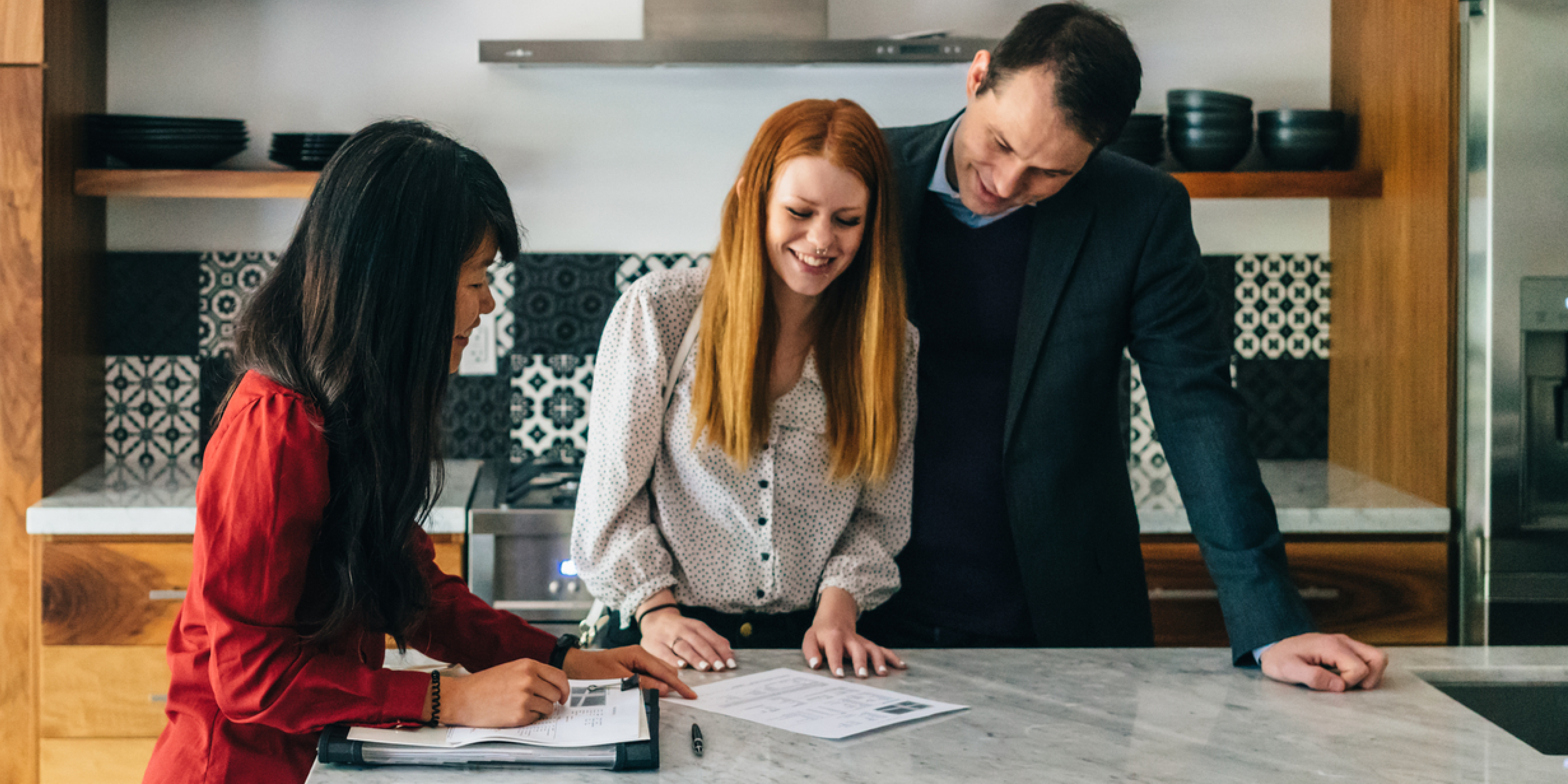 An image of three people with a man and a woman signing a lease for their new home in a kitchen. 