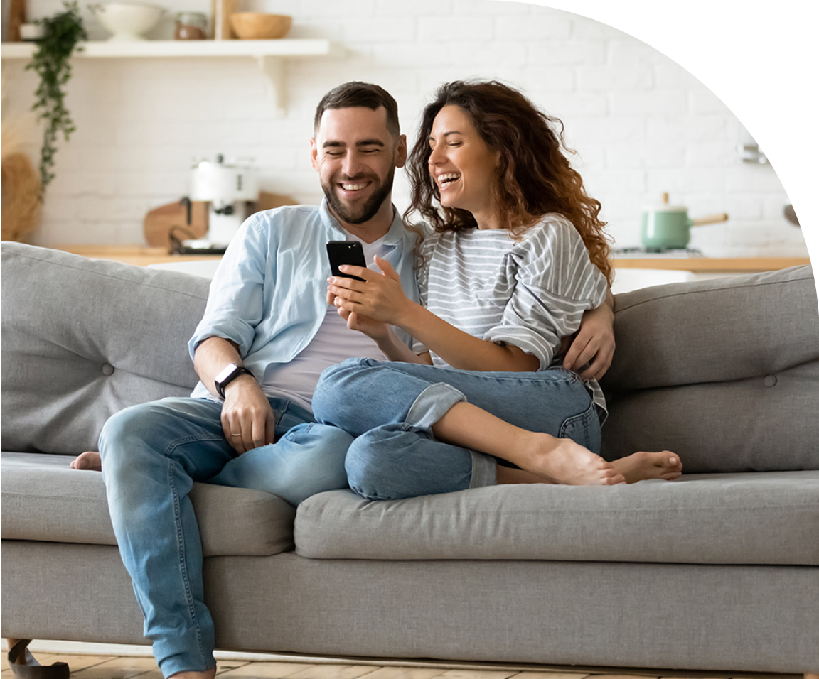 An image of a couple relaxing together on a grey couch, smiling at a phone one of them is holding