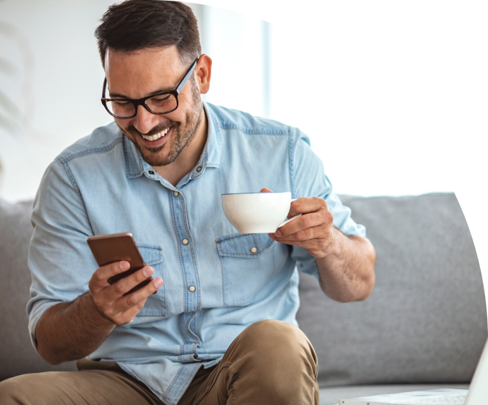 An image of a man holding a cup of coffee and sitting on a couch, smiling at his phone