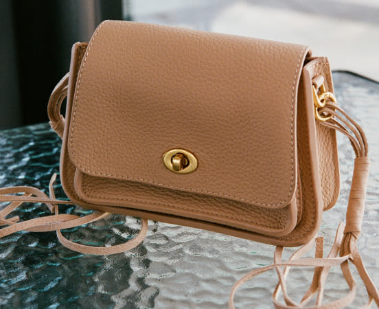 Tan leather crossbody purse with gold turnlock clasp and adjustable strap on dark green marble surface