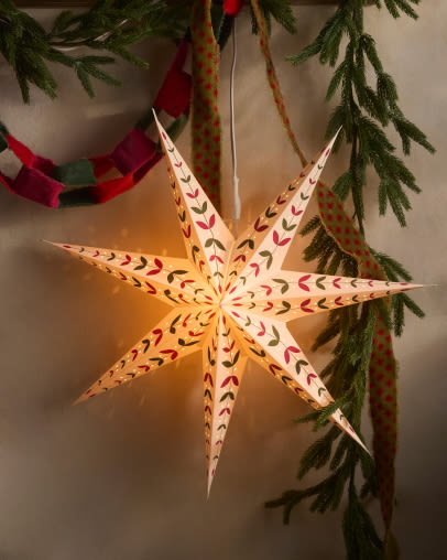 Illuminated white paper star ornament with red and green decorative patterns hanging from evergreen branches with holiday decorations.