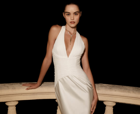 Woman in elegant white halter dress with deep V-neck and pendant necklace, seated on stone balustrade against black background