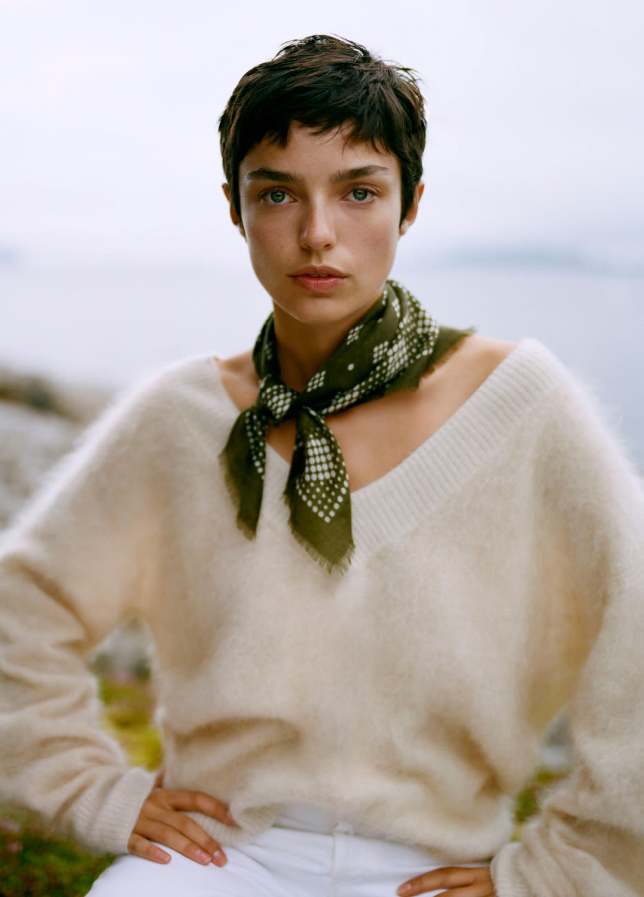 Woman with short dark hair wearing cream sweater and olive green patterned scarf, outdoors with blurred coastal background.