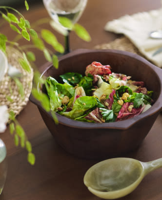 Fresh salad with mixed greens and toppings in a dark wooden bowl on a dining table with greenery and place settings