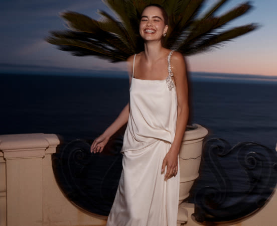Woman in white dress laughing with hair flowing on ocean balcony at dusk. Text reads BOOK AN APPOINTMENT.