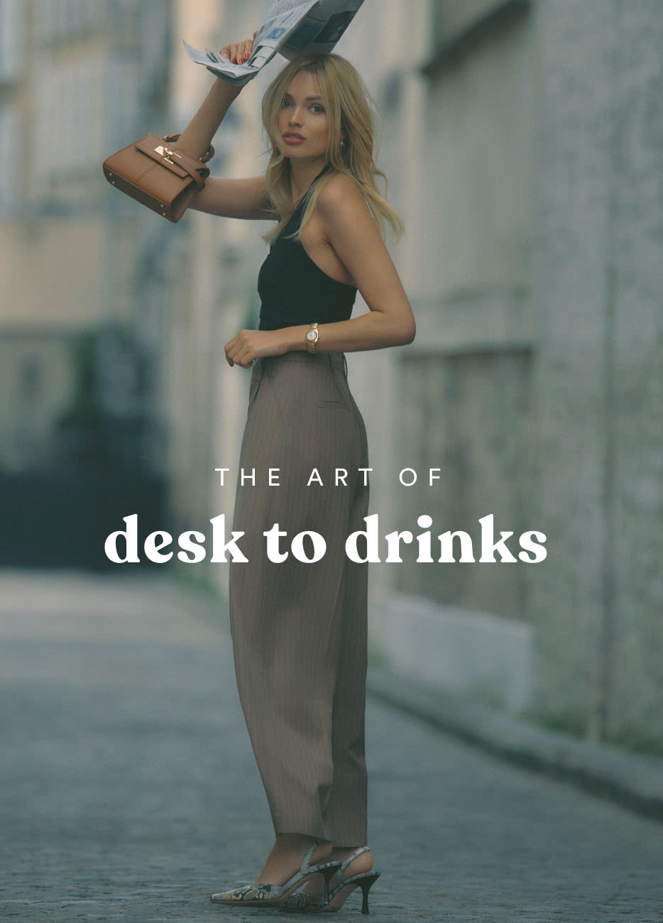 Woman in black top and gray pants holding magazine and purse with text THE ART OF desk to drinks on blurred city street.