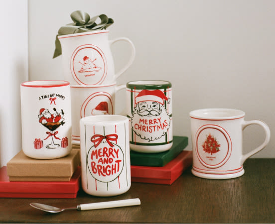 Collection of festive white mugs and candles with red Christmas designs including Santa, wine glass, and holiday text on books