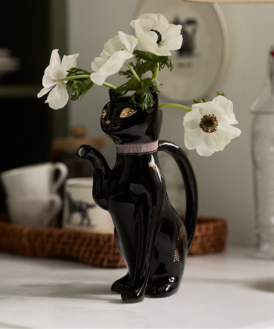 White anemone flowers in a black cat-shaped ceramic vase with gold eyes and pink collar on a white table.