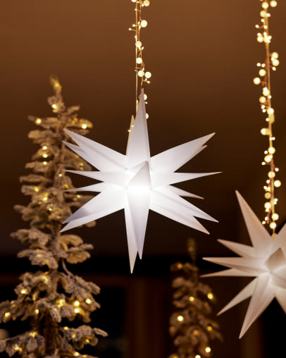 White illuminated star ornament hanging with golden Christmas tree and string lights in background against brown wall.