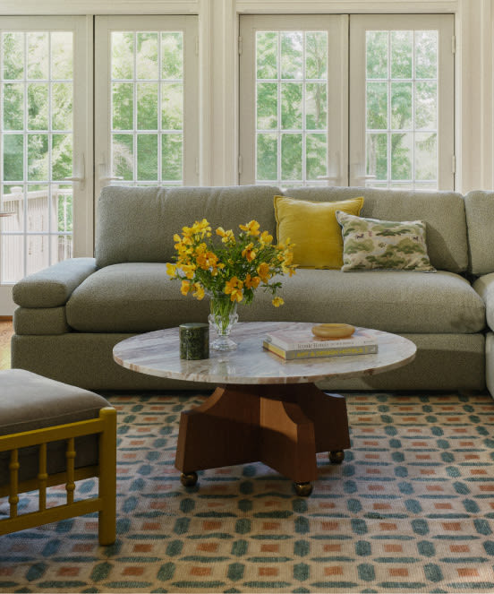 Gray sofa with yellow pillows behind round marble coffee table with yellow flowers in bright living room with large windows