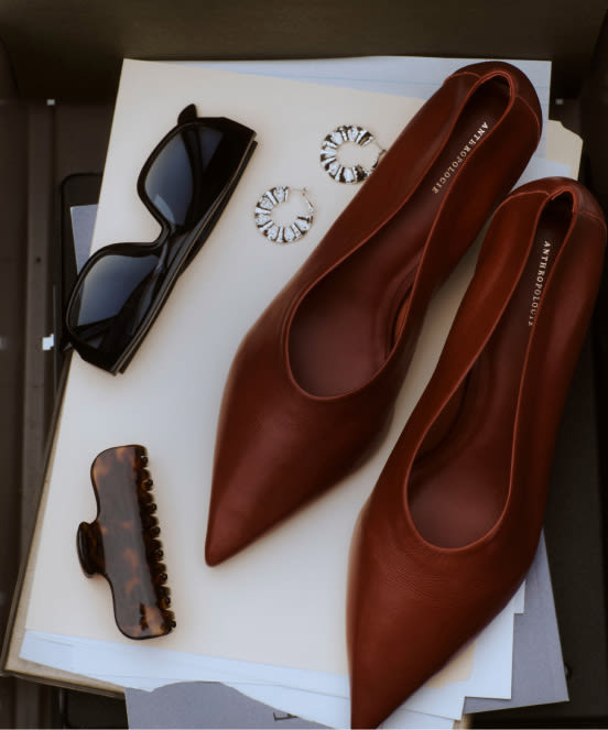 Brown leather pointed-toe flats on white papers with sunglasses, daisy earrings, and a brown hair clip arranged nearby.