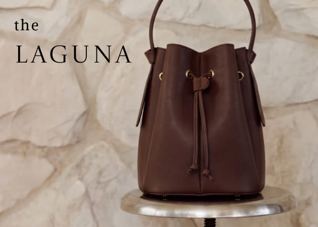 Brown leather drawstring bucket bag labeled "the LAGUNA" displayed on a marble pedestal against a white fabric backdrop.