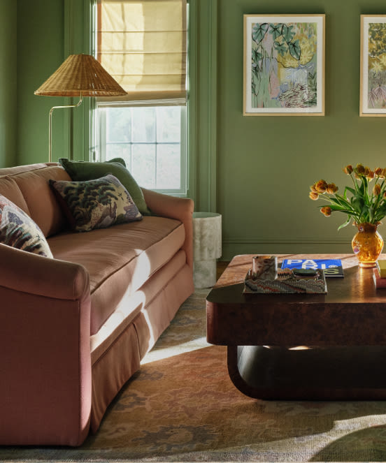 Cozy living room with sage green walls, pink velvet sofa, wicker lamp, framed artwork, and yellow tulips in amber vase.