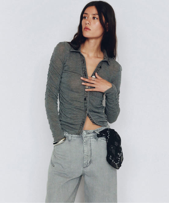 Woman in gray button-up shirt and light gray jeans looking to the side against a white background.