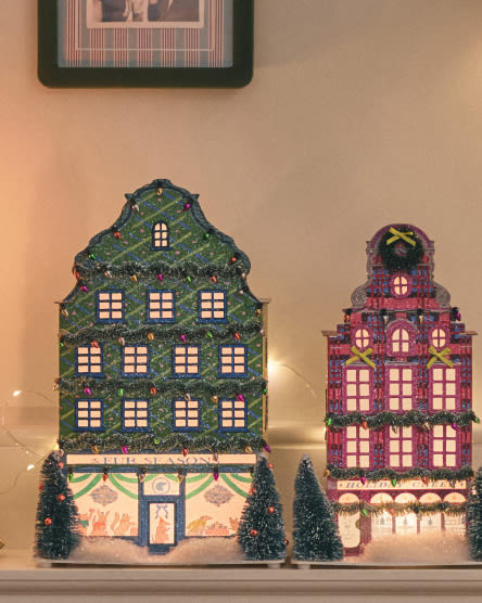 Two illuminated decorative house buildings with lit windows, one green and one purple, displayed on a shelf with small trees.