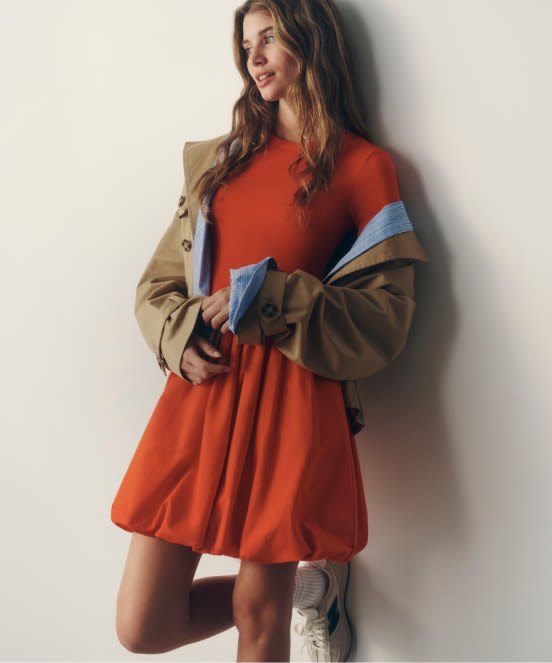 Woman in orange dress holding tan jacket with blue shirt, wearing white sneakers, standing against white wall.