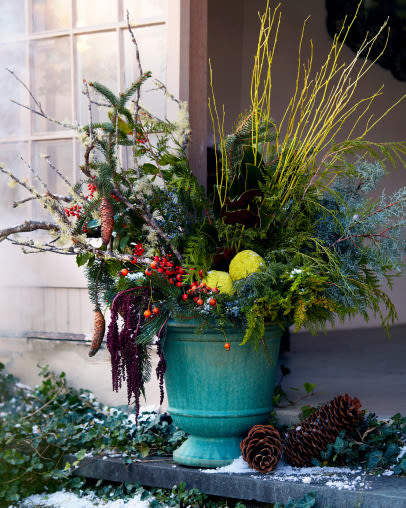Festive winter arrangement in turquoise urn with evergreen branches, red berries, yellow twigs, and pine cones on snowy doorstep.