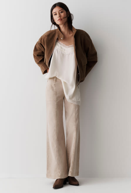 Woman wearing brown bomber jacket over white lace-trimmed camisole with beige wide-leg linen pants and brown loafers.