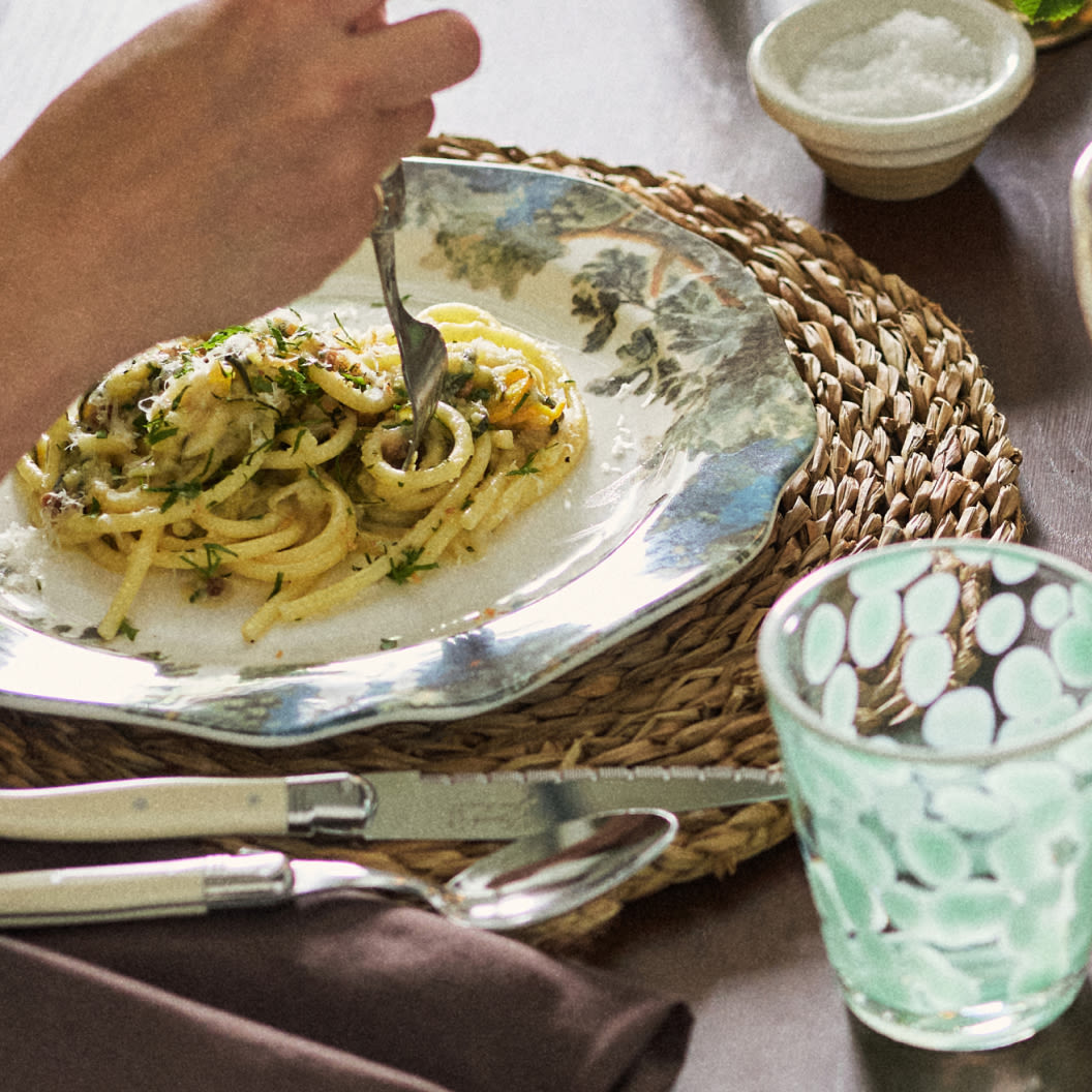 Hand twirling pasta with herbs on decorative plate over woven placemat, with mint green glass and utensils nearby.