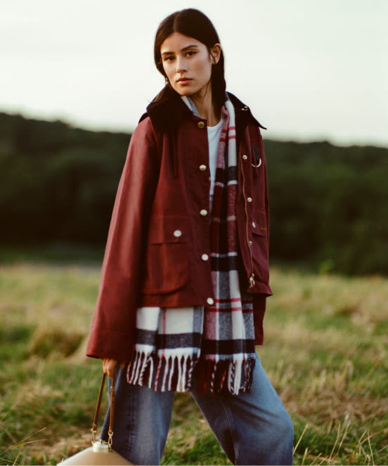 Woman in burgundy jacket with plaid scarf standing in grassy field with hills in background