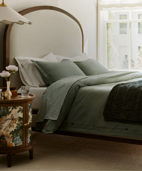 Bedroom with wooden arched headboard, sage green bedding, white pillows, round nightstand with lamp and flowers, bright window.