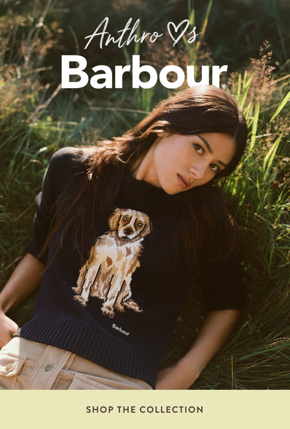 Woman in black Barbour sweater with dog graphic lying in grass. Text reads Anthro loves Barbour, Shop the Collection.