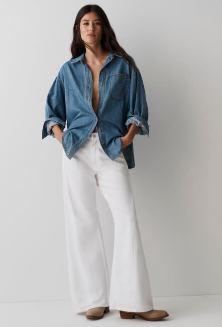 Woman wearing oversized blue denim shirt unbuttoned over white wide-leg pants with brown shoes against gray background