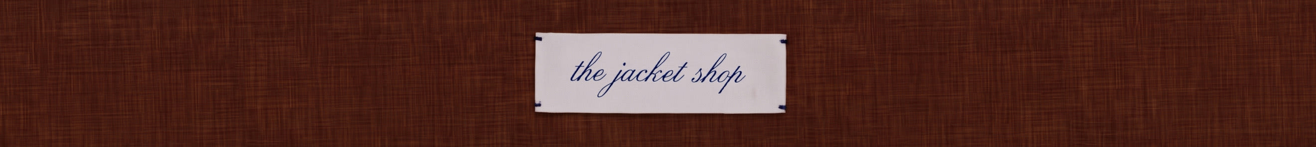 White rectangular sign with blue cursive text reading "the jacket shop" on a dark brown woven textured background