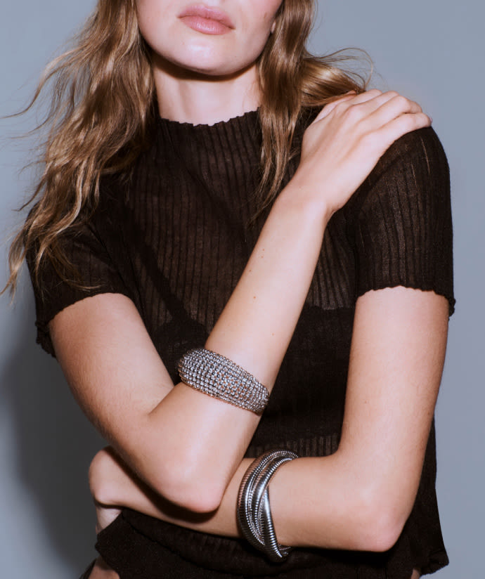 Woman with long brown hair wearing black ribbed top and silver rhinestone bracelets on both wrists against gray background.