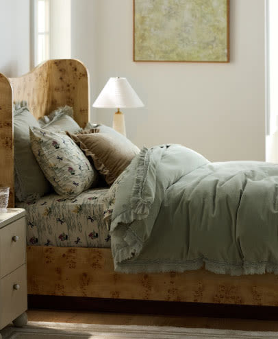 Cozy bedroom with wooden bed frame, sage green bedding, floral pillows, white nightstand, table lamp, and framed art.