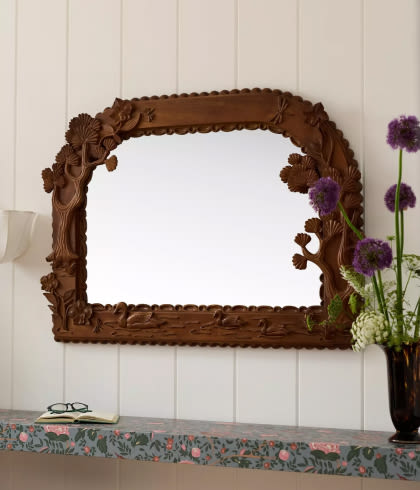 Ornate brown carved wooden mirror frame with floral details on white paneled wall above floral patterned shelf with purple flowers.